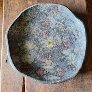 Puede incluir: Un gran bol de servir de cerámica gris con una superficie texturizada y moteada. El bol tiene una forma ligeramente irregular y presenta una pequeña pegatina dorada con el texto "Hirsch Art" y "Handmade".