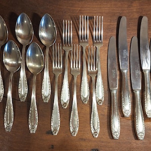 May include: A set of antique silverware, comprising spoons, forks, and knives, displayed on a wooden surface. The cutlery features a decorative floral design on the handles. The spoons, forks, and knives are made of a shiny, silver-coloured metal.