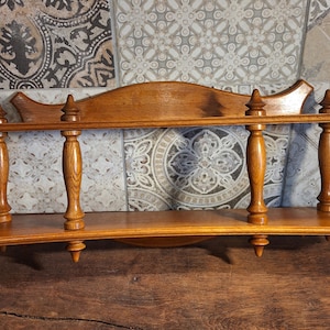 May include: A two-tiered wooden shelf in a warm, medium-brown hue. It features decorative turned supports and a curved design. The top shelf has a scalloped edge. The shelf is designed for wall mounting.