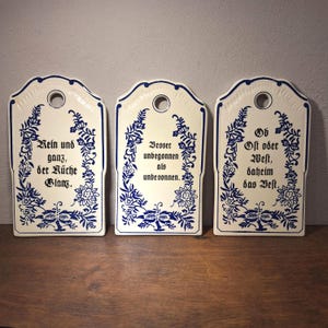 Könnte beinhalten: Drei cremefarbene Keramikplatten mit blauen Blumenmustern und deutschem Text. Jede Platte hat oben ein Loch zum Aufhängen. Der Text enthält Sätze wie "Rein und ganz, der Küche Glanz" und "Ob Ost oder West, daheim das Vest."