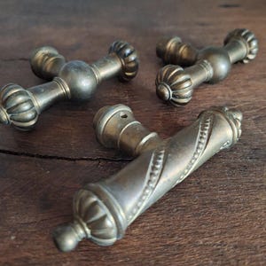Op de afbeelding: Drie antieke messing deurgrepen. Twee grepen hebben een kruisvorm met sierlijke details. De derde greep is een lang, gedraaid ontwerp met een decoratieve knop aan elk uiteinde.