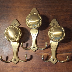 Könnte beinhalten: Drei Wandhaken aus Messing mit einem Blumendesign auf der runden Oberseite. Jeder Haken hat ein Doppelhakendesign und eine dekorative Rückplatte. Die Haken sind auf einer dunklen Holzoberfläche montiert.