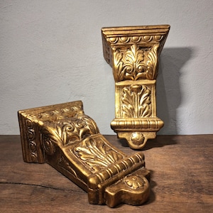 May include: Two gold-coloured wall corbels featuring intricate carved leaf patterns and decorative moulding. Each piece has a flat top surface, a curved body with detailed foliage relief, and a small circular floral accent at the bottom base.