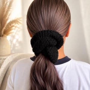 May include: A black knitted hair scrunchie with a textured, ribbed surface. The accessory is circular in shape and features a thick, yarn-like construction that gathers hair into a ponytail, providing a secure hold with a soft, fabric-based finish.