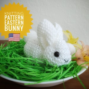 May include: White knitted Easter bunny sitting in a bed of green grass. The bunny has black eyes and a small pink nose. The bunny is sitting in a white bowl.