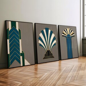 May include: Three framed art prints with Art Deco designs. The prints feature geometric shapes in teal, gold, and white against a gray background. The frames are black, and the artwork is displayed on a wooden floor.