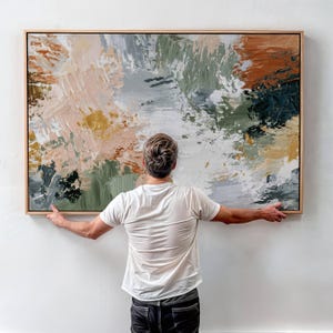 May include: A large rectangular abstract painting in a light wood frame featuring thick, textured brushstrokes in shades of sage green, burnt orange, cream, white, and dark charcoal gray, displayed on a plain white wall surface.