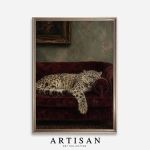 May include: A framed wall art print featuring a snow leopard resting on a deep burgundy velvet tufted sofa. The leopard is positioned with its head on a round bolster pillow. Above the sofa, a portion of a dark framed painting is visible.