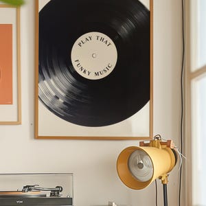 May include: A framed print of a black vinyl record with the text "Play That Funky Music" in white. The print is hanging on a white wall next to a record player and a yellow lamp.