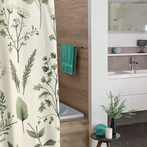 Puede incluir: Una cortina de ducha blanca con estampado bot&aacute;nico verde cuelga en un ba&ntilde;o. Una toalla azul verdosa cuelga de un toallero y una toalla de mano azul verdosa se encuentra sobre una peque&ntilde;a mesa auxiliar de madera con un jarr&oacute;n negro y una vela blanca.