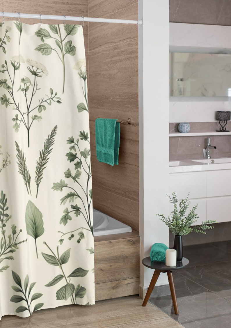 Puede incluir: Una cortina de ducha blanca con estampado bot&aacute;nico verde cuelga en un ba&ntilde;o. Una toalla azul verdosa cuelga de un toallero y una toalla de mano azul verdosa se encuentra sobre una peque&ntilde;a mesa auxiliar de madera con un jarr&oacute;n negro y una vela blanca.