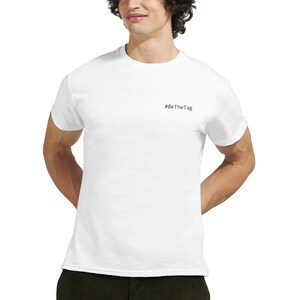 Puede incluir: Una camiseta blanca de cuello redondo y manga corta. El texto "#BeTheTag" está impreso en negro en la parte superior del pecho. La camiseta está hecha de un material suave.