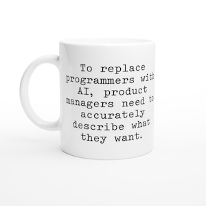 Könnte beinhalten: Eine weiße Keramiktasse mit schwarzem Text, der lautet: "Um Programmierer durch KI zu ersetzen, müssen Produktmanager genau beschreiben, was sie wollen."
