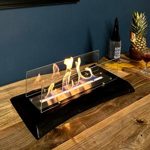 Può includere: Un moderno caminetto nero e in vetro con fiamme, posto su un tavolo di legno. Un ritratto di leopardo incorniciato è appeso a una parete blu scuro. Una bottiglia, un ananas e un bicchiere di vino sono anche sul tavolo.