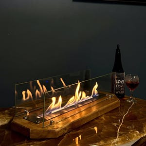 Puede incluir: Una chimenea rectangular con base de madera y paneles de vidrio, que muestra una llama vibrante. Una botella oscura con las palabras "LOVE LIFE WATER" y una copa de vino tinto están junto a la chimenea. El conjunto está sobre una superficie similar al mármol.