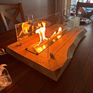 以下が含まれることがあります： 生木端の長方形の木製卓上暖炉。暖炉はガラス製のエンクロージャーと内部で燃える炎を備えています。木材は暖かく赤褐色の色をしています。暖炉は木製のテーブルの上にあります。