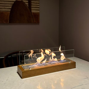 以下が含まれることがあります： 木製の台座とガラスパネルが付いた卓上暖炉。暖炉は点火されており、炎が燃えています。