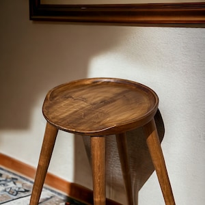 Könnte beinhalten: Ein Hocker aus Holz mit einer runden, leicht konkaven Oberseite und drei Beinen. Der Hocker ist aus dunkelbraunem Holz gefertigt und hat eine natürliche, rustikale Oberfläche.