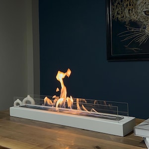 Op de afbeelding: Witte tafelhaard met een glazen schild en een realistisch vlammeneffect. De haard staat op een houten tafel.
