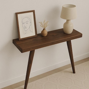 May include: A dark brown wooden console table with a minimalist design. It features a framed line drawing, a small vase with dried flowers, and a beige table lamp. The table has a rectangular top and angled legs.