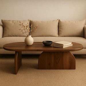 May include: A dark brown, oval-shaped coffee table with a curved base and two supporting legs. A small, round, black bowl and two books sit on the table. A beige vase with dried flowers is also on the table. A beige sofa is in the background.
