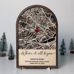 Puede incluir: Una pieza de arte de mapa de madera con un diseño de mapa de la ciudad cortado con láser, con un corazón rojo que marca una ubicación. El texto "Where it all began" está inscrito, junto con nombres y una fecha. La pieza se muestra sobre un soporte, con árboles en miniatura en el fondo.