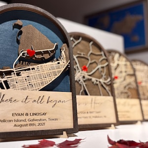 May include: An arched wooden map display featuring a laser-cut street grid of Pelican Island, Galveston, Texas, set against a dark blue background. A small red heart marks a location. Text reads: Where it all began, EVAN & LINDSAY, August 8th, 2021.