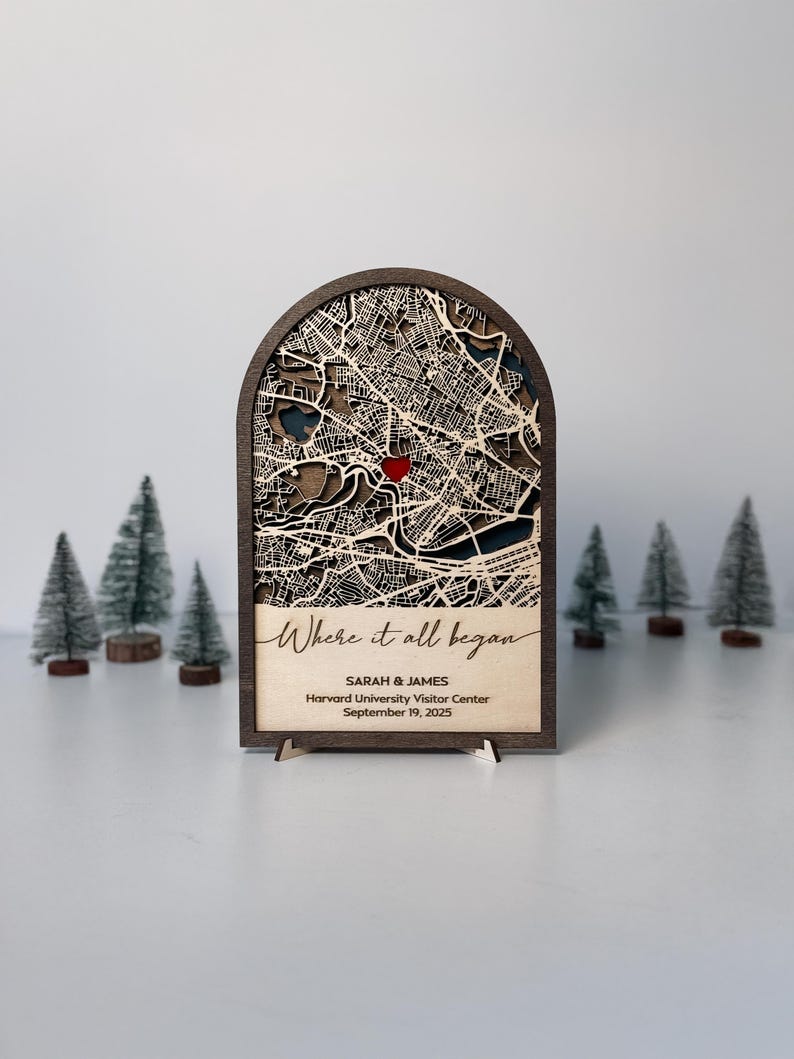 May include: A wooden map art piece with a laser-cut city map design, featuring a red heart marking a location. The text "Where it all began" is inscribed, along with names and a date. The piece is displayed on a stand, with miniature trees in the background.