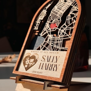 May include: A wooden layered map of a city with a red heart marking a location. The frame includes a photo of a couple and the text "WHERE IT ALL BEGAN" and "SALLY HARRY JUNE 20, 1977". The piece is displayed on a wooden stand.