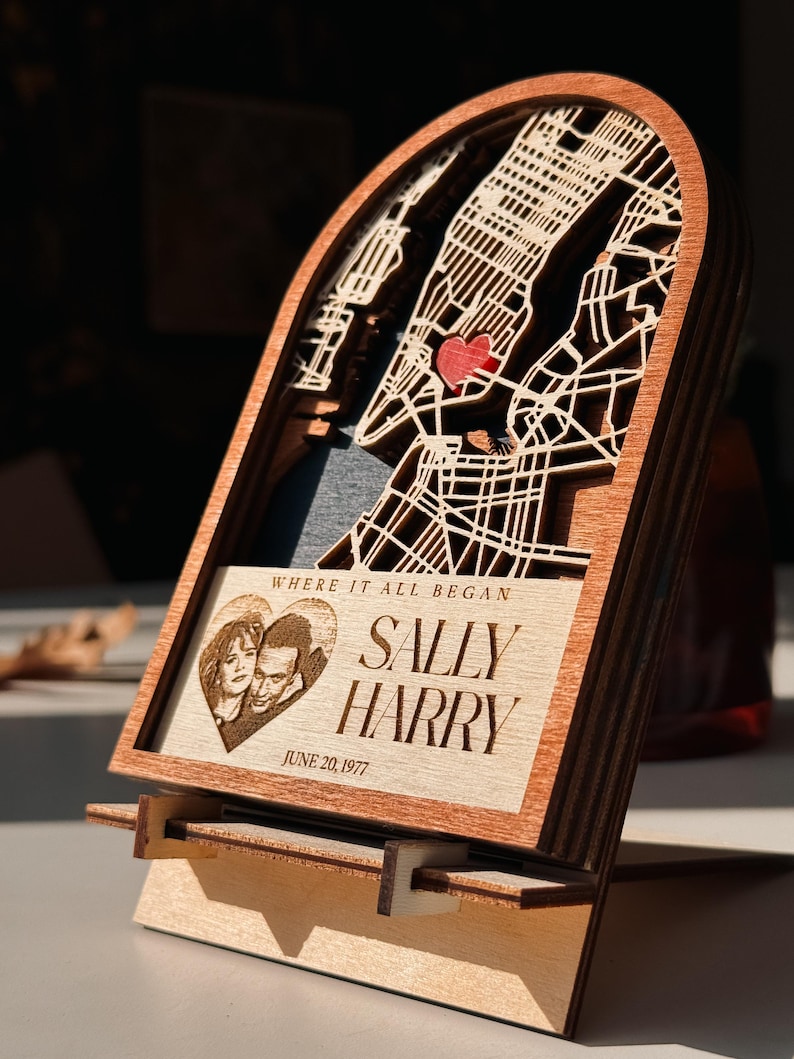 May include: A wooden layered map of a city with a red heart marking a location. The frame includes a photo of a couple and the text "WHERE IT ALL BEGAN" and "SALLY HARRY JUNE 20, 1977". The piece is displayed on a wooden stand.