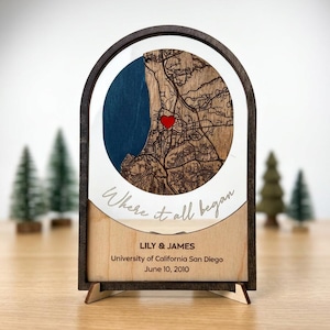 May include: A wooden map art piece with a dark brown frame and a light wood base. The map features a blue section representing water and a detailed brown land area with a red heart. Text includes "Where it all began," and names and a date.