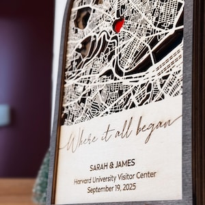May include: A wooden laser-cut map with a red heart marking a location. The map is framed and features the text "Where it all began," along with names, a location, and a date. The frame is dark brown, and the map is light-colored.