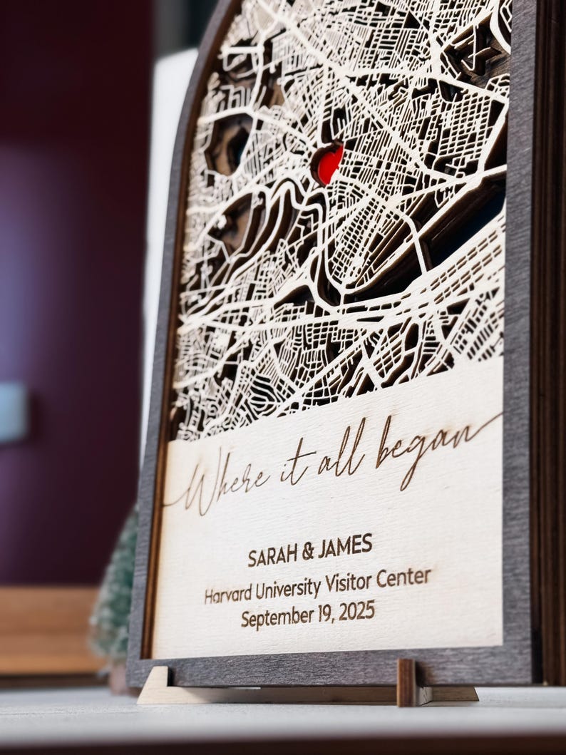 May include: A wooden laser-cut map with a red heart marking a location. The map is framed and features the text "Where it all began," along with names, a location, and a date. The frame is dark brown, and the map is light-colored.