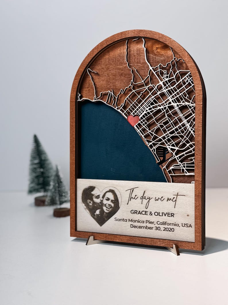 May include: Wooden map art piece with a laser-cut city map, a heart-shaped photo, and the text "The day we met." The map features a red heart marking a location. The piece includes the names Grace & Oliver and the date December 30, 2020.
