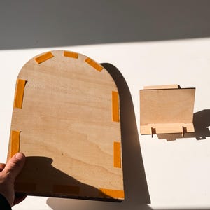 May include: Two unfinished wooden pieces. One is a large, arched rectangle with orange adhesive tape on the edges. The other is a small, rectangular stand. Both are made of light-colored wood and are set against a white background.