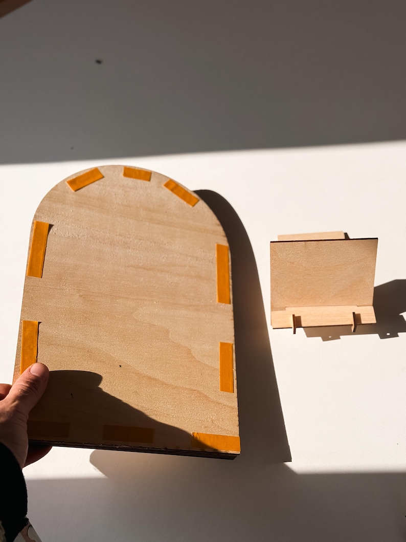 May include: Two unfinished wooden pieces. One is a large, arched rectangle with orange adhesive tape on the edges. The other is a small, rectangular stand. Both are made of light-colored wood and are set against a white background.