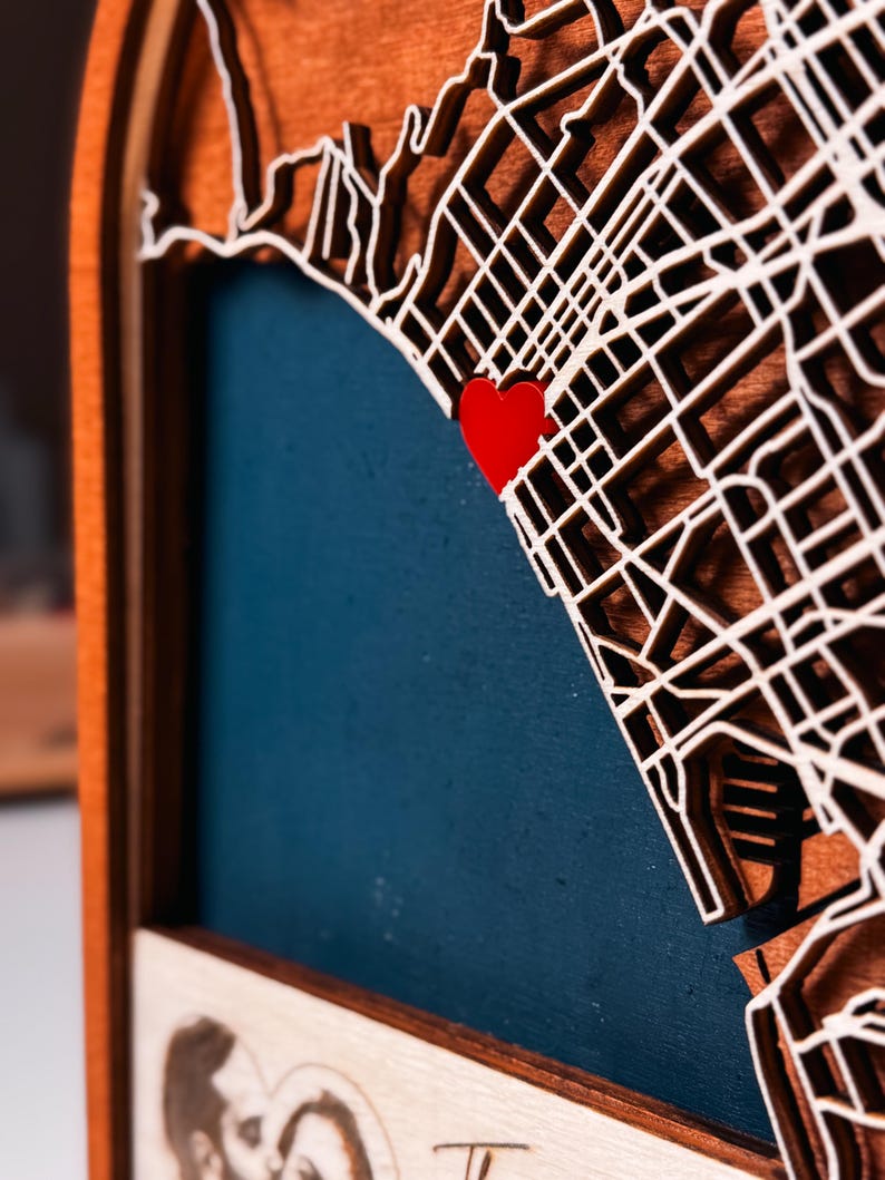 May include: A layered wooden map with a red heart marking a location. The map features a detailed street grid and coastline, set against a blue background. The frame is made of dark wood.