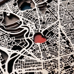 May include: A layered wooden map with intricate street and building cutouts. The map features a red heart-shaped cutout, possibly indicating a special location. The wood has a natural, light color, and the design is detailed and precise.