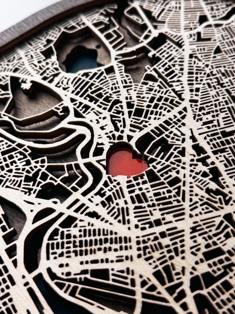 May include: A layered wooden map with intricate street and building cutouts. The map features a red heart-shaped cutout, possibly indicating a special location. The wood has a natural, light color, and the design is detailed and precise.
