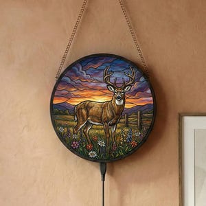 Op de afbeelding: Ronde wandkunst met een hert in een veld met een zonsondergang op de achtergrond. Het kunstwerk is ingelijst in een zwart frame en hangt aan een ketting. Een zwart snoer met een aan/uit-schakelaar hangt eronder.