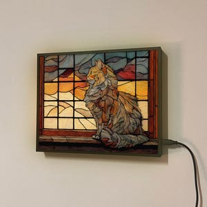 Peut inclure: Une fenêtre en vitrail représentant un chat assis sur le rebord, regardant un paysage de coucher de soleil. La fenêtre est encadrée de bois foncé et est équipée d'un cordon avec un interrupteur pour allumer une lumière.