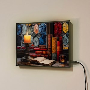 May include: A framed wall art piece with a stained glass window design featuring a lit candle, books, and a small jar. The image is set against a dark wood background.