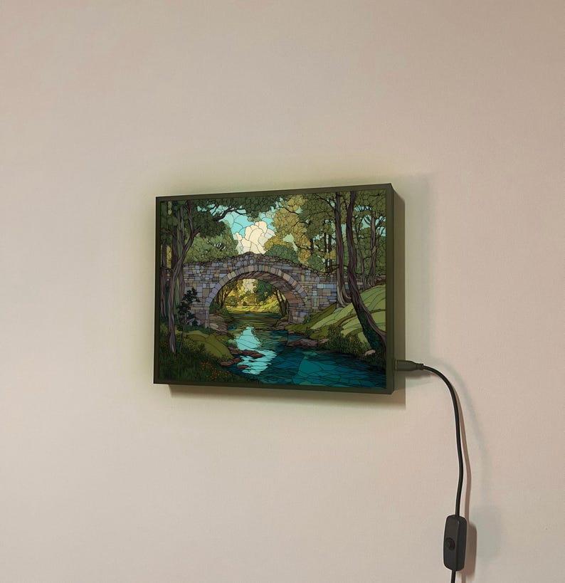May include: A framed picture showing a stone bridge over a river, with trees on either side. The artwork uses greens, blues, and browns. The frame is dark green, and a black power cord with a switch is attached.
