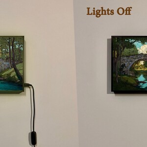 May include: A framed landscape artwork showing a stone bridge over a river, surrounded by trees. The image is displayed with the lights on and off, showing the illuminated and non-illuminated versions. The frame is dark.