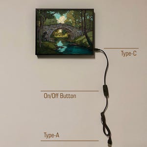 May include: A framed artwork showing a stone bridge over a river in a forest. It has a Type-C and Type-A connector, and an On/Off button. The artwork is mounted on a white wall. The artwork is connected to a power source.