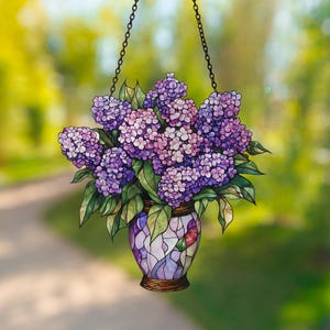 May include: A stained glass window hanging with a purple and green floral design. The flowers are in a vase with a mosaic pattern. The background is blurred.