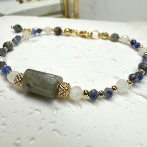 May include: A beaded bracelet featuring a central cylindrical gray stone bead flanked by two gold-toned textured spacers. The design includes alternating faceted blue, gray, and white beads interspersed with small gold-toned metal spheres.
