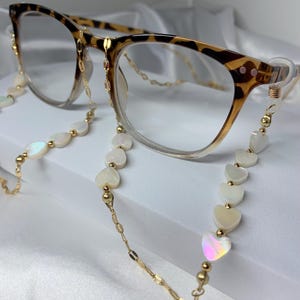 Puede incluir: Una cadena dorada con cuentas blancas en forma de corazón unidas a un par de gafas de carey.