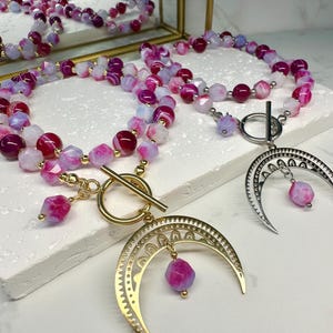 May include: Necklaces with pink, purple, and white beads. One necklace has a gold crescent moon pendant, and the other has a silver crescent moon pendant. The necklaces are displayed on a white surface.