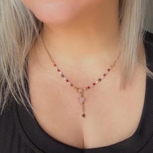 Könnte beinhalten: Eine goldene Halskette mit einem zentralen rosa herzförmigen Anhänger und einer kleinen hängenden Perle. Die Kette ist mit einer Reihe abwechselnder kleiner roter, weißer und dunkler Perlen verziert und mit einem runden Verschluss versehen.